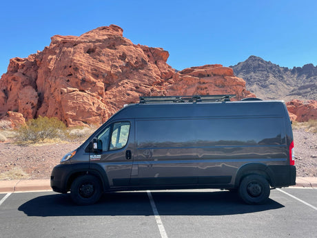 Dark gray Ram ProMaster van (potential RV rental) parked near striking red rock formations under a clear blue sky, ideal for desert travel.
