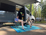 Two people relax in chairs outside a grey camper van, an RV rental, under an awning at a wooded campsite. Perfect outdoor getaway.