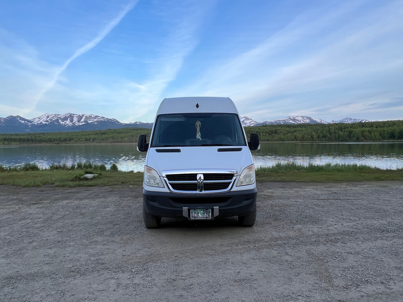 This RV rental van is parked on gravel by an Alaskan lake, with snow-capped mountains and blue sky.