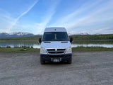 This RV rental van is parked on gravel by an Alaskan lake, with snow-capped mountains and blue sky.