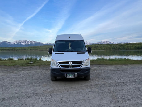 This RV rental van is parked on gravel by an Alaskan lake, with snow-capped mountains and blue sky.