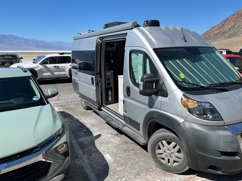  and swivel chairs.|A silver rv rental camper van is parked at a sunny desert campsite with a picnic table and red rock mesas under a clear blue sky.|Woman gives a thumbs-up from her chair next to an RV rental camper van in a vast desert landscape with mountains.|Silver RV rental parked in a sunny desert landscape with mountains in the background. Side door open — photo 5