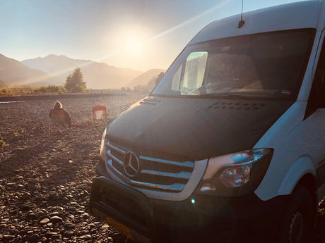 Adventure awaits! An RV rental Sprinter van parked on a rocky riverbed at sunset, with mountains & a person camping.