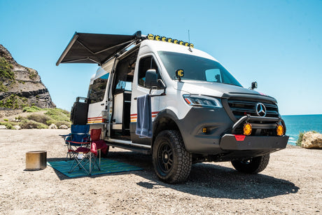 Customized Sprinter campervan with awning & camp chairs by the beach. Ideal off-road rv rental for adventure.