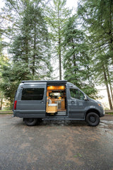 Custom grey camper van RV rental in a lush forest. Side door open, revealing a warm, functional interior kitchen & living space.