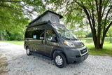 A dark grey Ram ProMaster camper van RV rental with a pop-top roof, parked on a gravel path near a scenic lake.