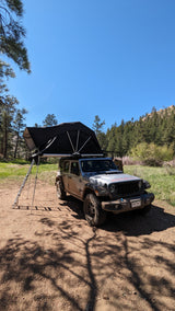 A silver RV rental Jeep with a rooftop tent deployed in a sunny, forested area for wilderness camping.