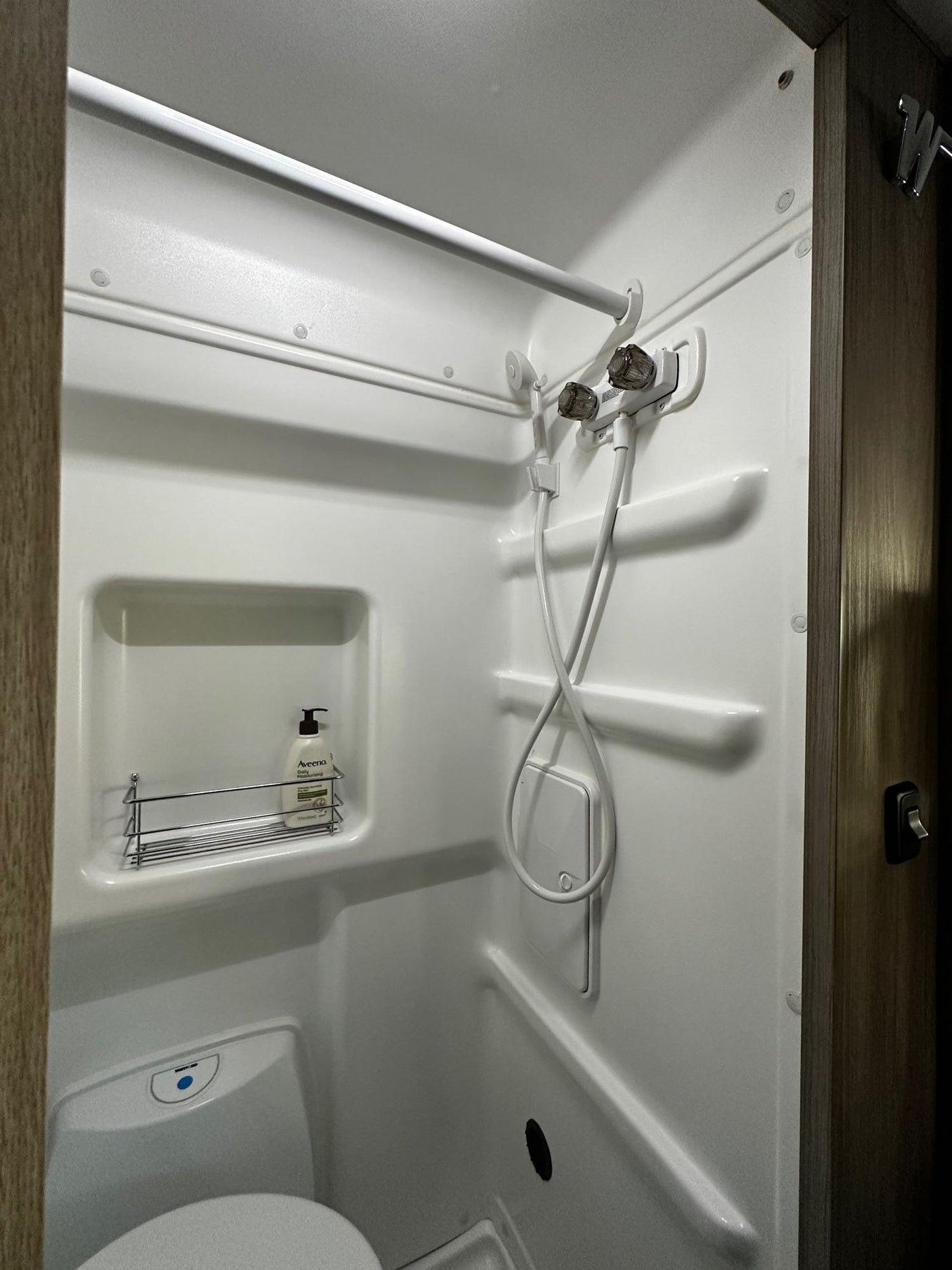 A compact, white RV rental bathroom, showing a built-in shower, toilet, and a wall shelf with toiletries.