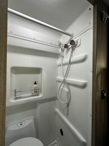 A compact, white RV rental bathroom, showing a built-in shower, toilet, and a wall shelf with toiletries.