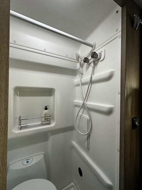 A compact, white RV rental bathroom, showing a built-in shower, toilet, and a wall shelf with toiletries.