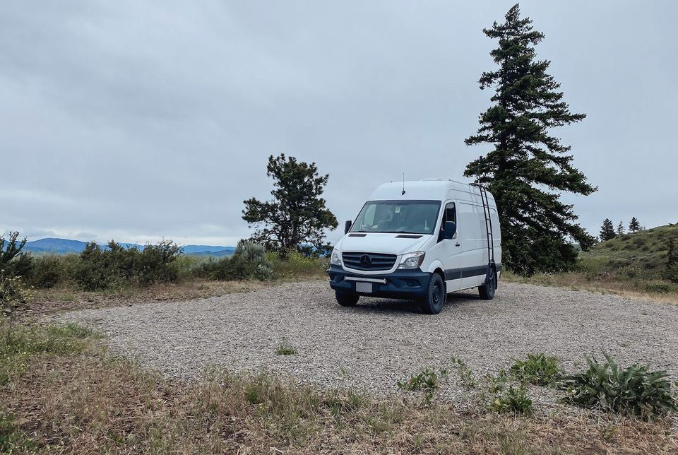 Explore the outdoors! This white Mercedes Sprinter campervan, perfect for an rv rental adventure, is parked on a gravel spot with trees & mountains.