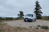 Explore the outdoors! This white Mercedes Sprinter campervan, perfect for an rv rental adventure, is parked on a gravel spot with trees & mountains.