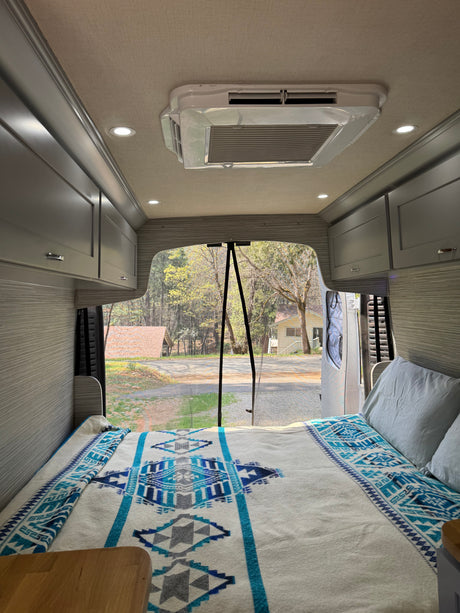 RV rental interior: A cozy bed with a blue patterned blanket, overhead cabinets, and open back doors reveal a wooded outdoor scene.