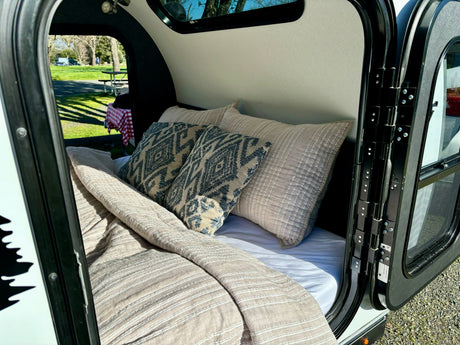 Cozy RV rental camper interior: A comfy bed with patterned pillows, looking out to a green campsite with a picnic table.