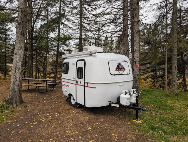 White Scamp travel trailer (RV rental) at a wooded campsite with a picnic table, perfect for your next camping adventure.