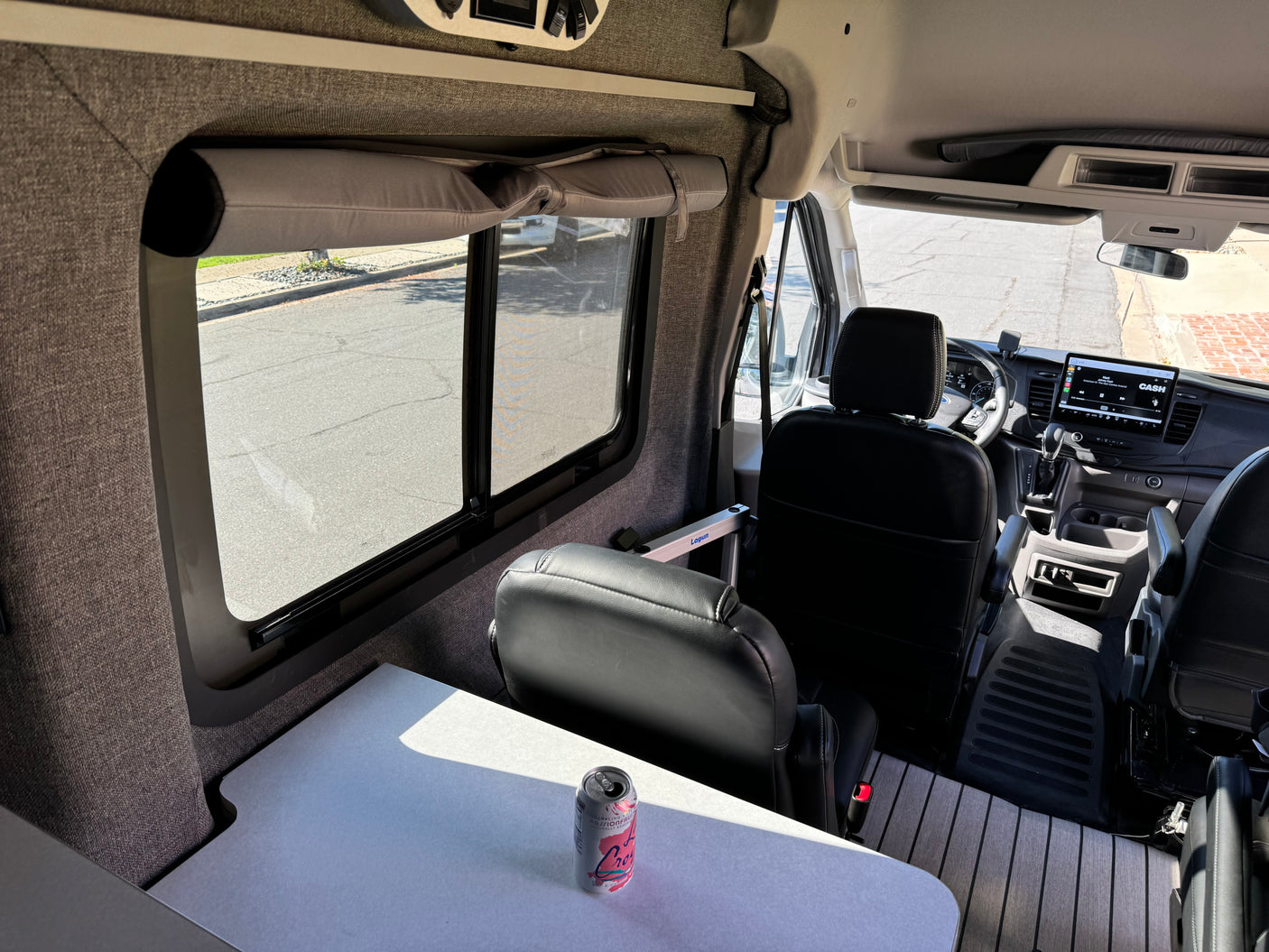 Cozy rv rental interior: table with LaCroix, large window with shade, front cabin, dashboard, & black leather seats.