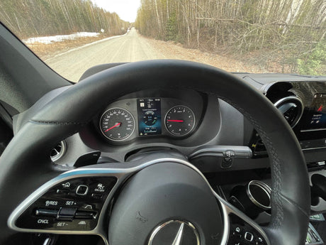 Inside an RV rental, dashboard shows 0 km/h and 318 mi range, with a gravel road and snowy nature ahead.