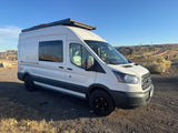 This white Ford Transit camper van, perfect for an RV rental adventure, is parked on a gravel lot under a clear blue sky.