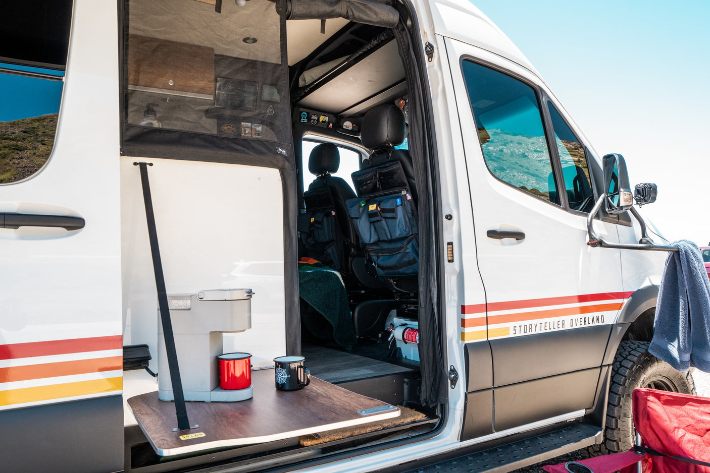 An open white RV rental with an outdoor coffee setup on a pull-out table, showing the interior and scenic view.