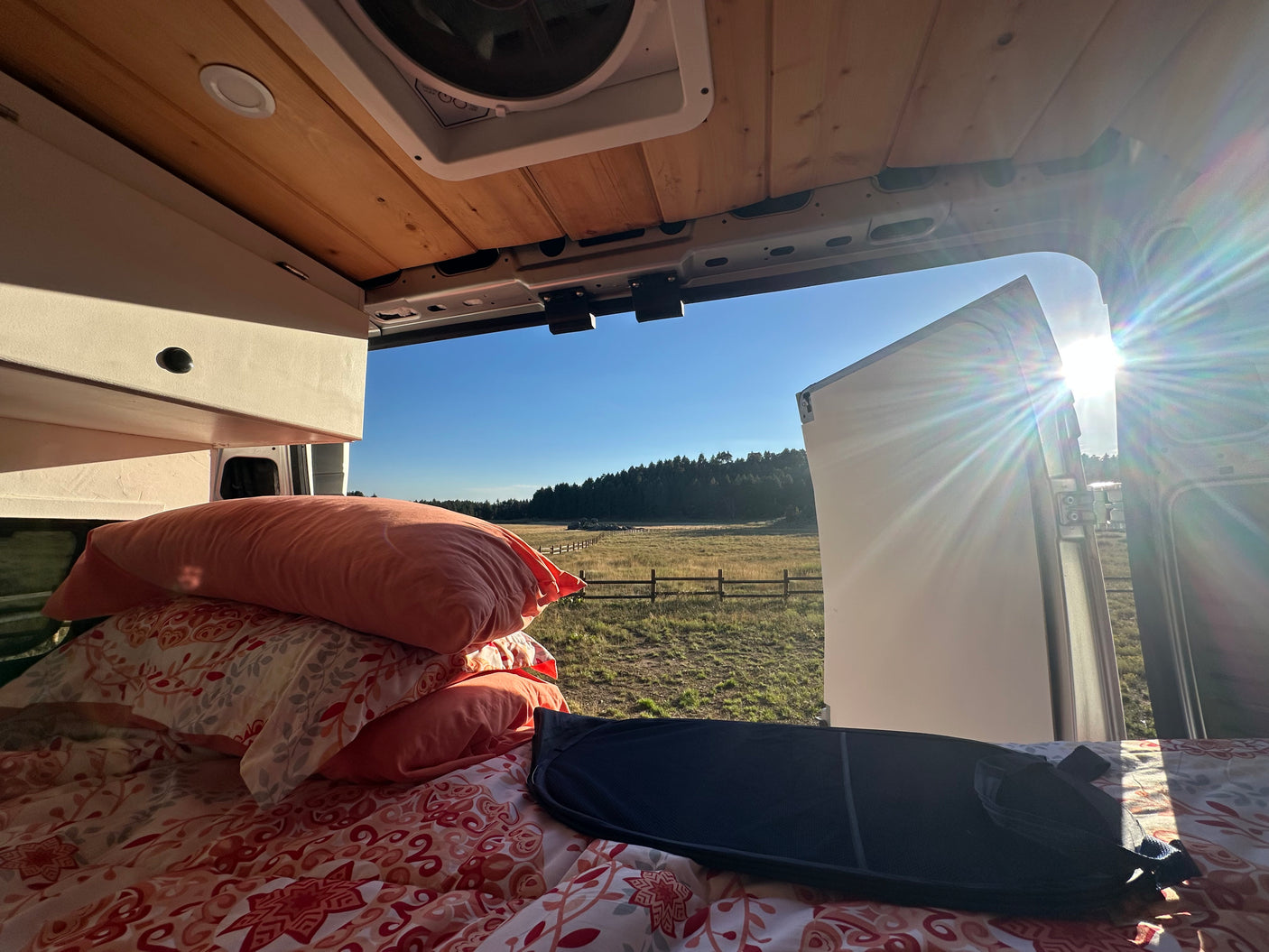 Inside an RV rental van: cozy bed, wooden ceiling, and a stunning, sun-drenched field view.