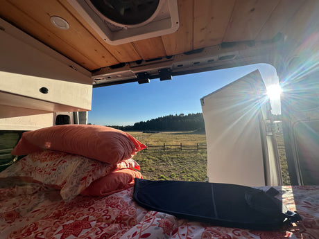 Inside an RV rental van: cozy bed, wooden ceiling, and a stunning, sun-drenched field view.