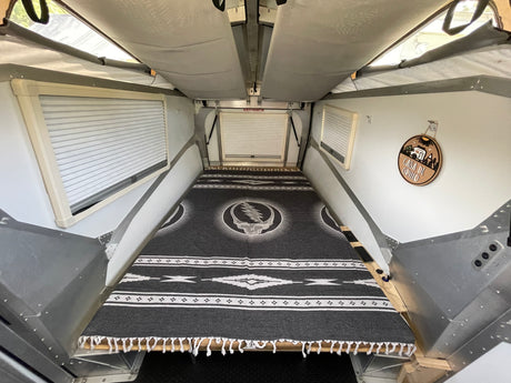 Inside a cozy pop-top RV rental, featuring a bed with a Grateful Dead blanket, window blinds, and an EXIT/SORTIE sign.