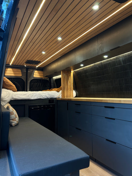 Explore this stylish RV rental's modern interior: a cozy bed, sleek dark kitchenette, wood ceiling with LED lighting.