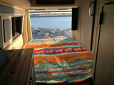 Cozy RV rental interior with a patterned bed overlooking a scenic ocean view from the rear, perfect for travel.