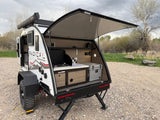 This RV rental is a white Radical Bushwacker teardrop trailer with its rear kitchen galley open, revealing a stove and cooler for outdoor camping.