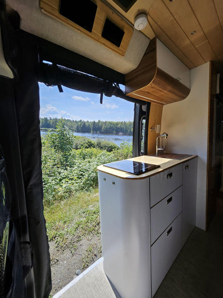 RV rental: Modern camper van kitchen with sink, induction cooktop, and a scenic river view outside.