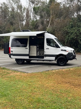 A white camper van with an extended awning and open side, parked in nature. Perfect for your next rv rental adventure!