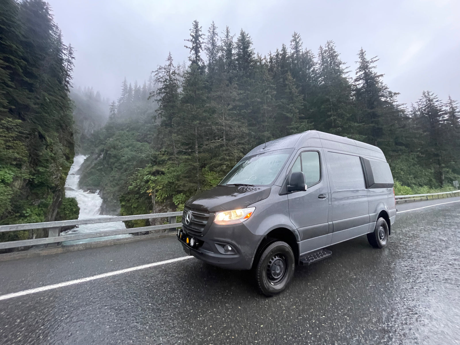 Grey Sprinter RV rental on a wet road beside a majestic waterfall and misty forest. Ideal for scenic adventures.