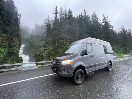 Grey Sprinter RV rental on a wet road beside a majestic waterfall and misty forest. Ideal for scenic adventures.