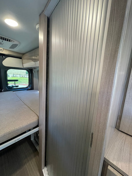 VANN camper van interior: an RV rental with a cozy bed, window view, and modern slatted door and wood-grain trim.