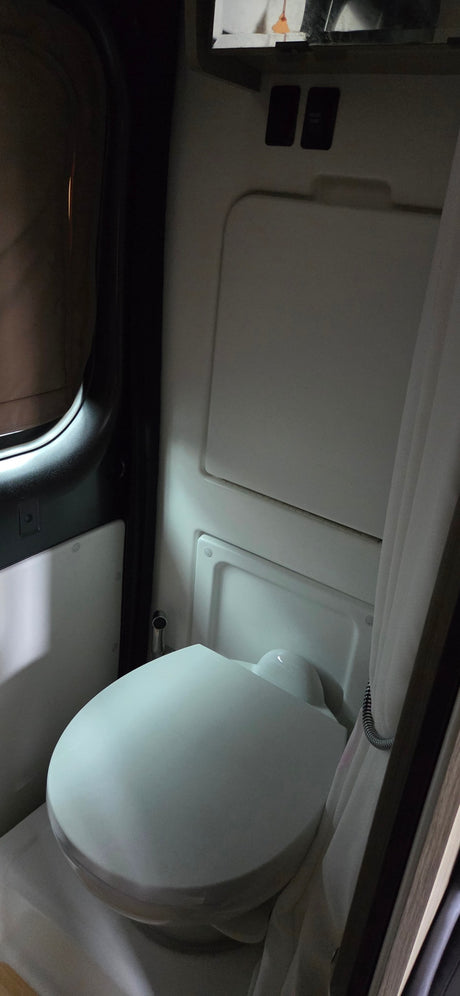 Inside a compact RV rental bathroom: a white toilet with closed lid, a shower curtain, and switches, including 'WATER PUMP'.