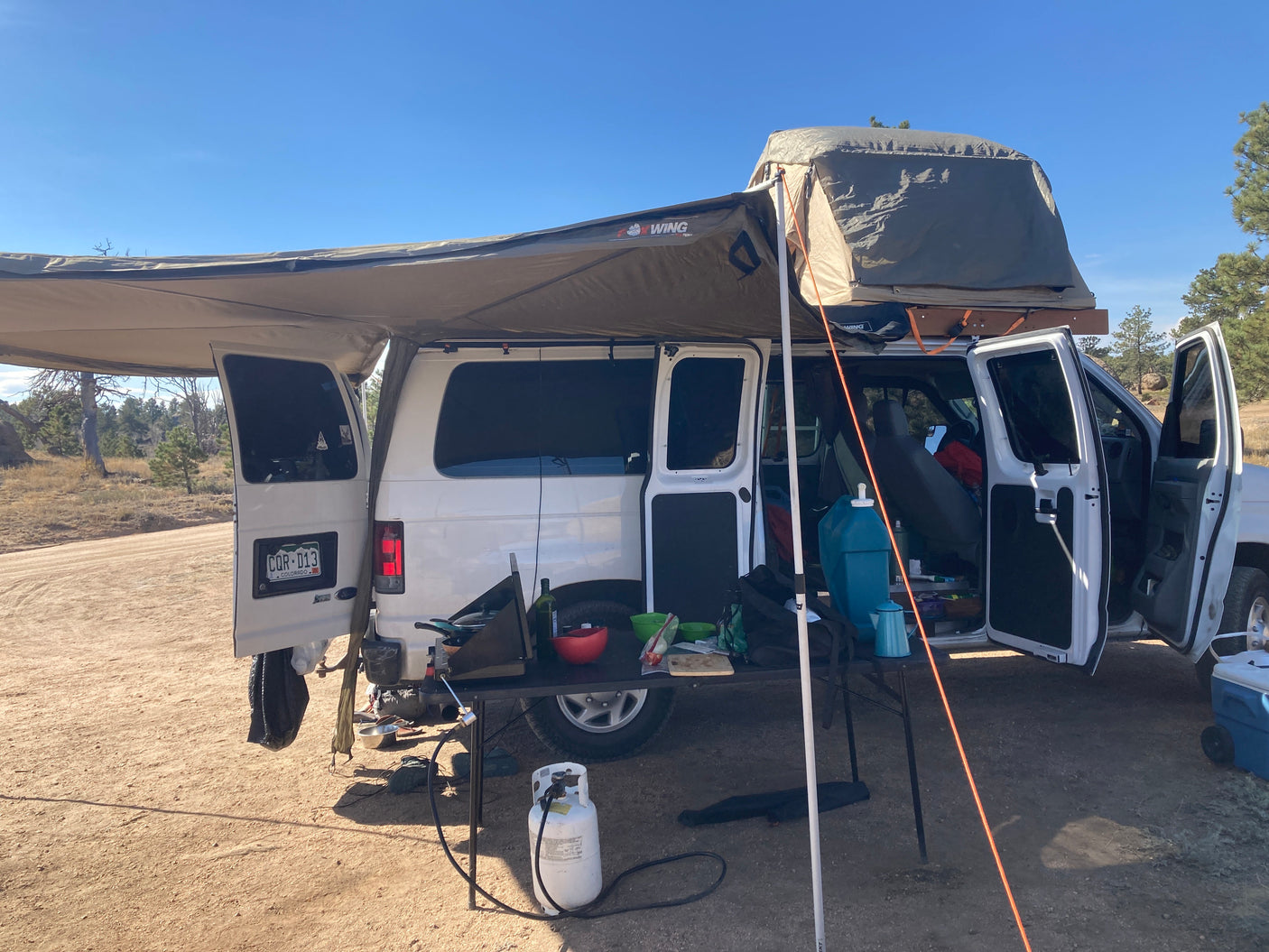 RV rental transformed: White camper van with large awning, rooftop tent, and outdoor kitchen setup at a sunny campsite.