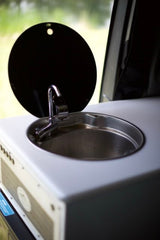 Inside an RV rental: a compact kitchen sink with a chrome faucet, the black circular lid is open. A practical mobile amenity.