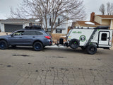 Blue Subaru Outback with roof rack towing a white, green NoBo camper on cracked asphalt. Ideal RV rental adventure setup!