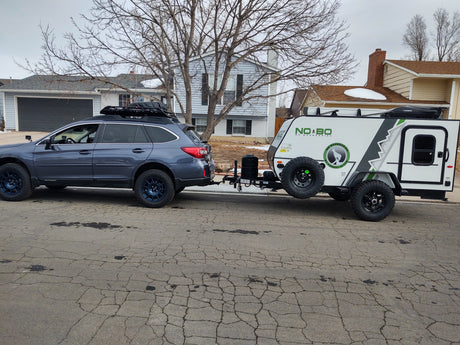 Blue Subaru Outback with roof rack towing a white, green NoBo camper on cracked asphalt. Ideal RV rental adventure setup!
