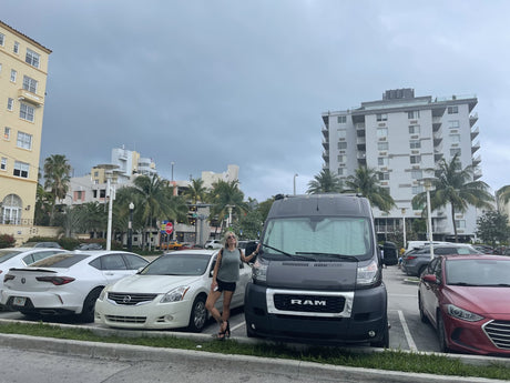 A woman stands by her grey RV rental camper van in a city parking lot, surrounded by buildings and palm trees.