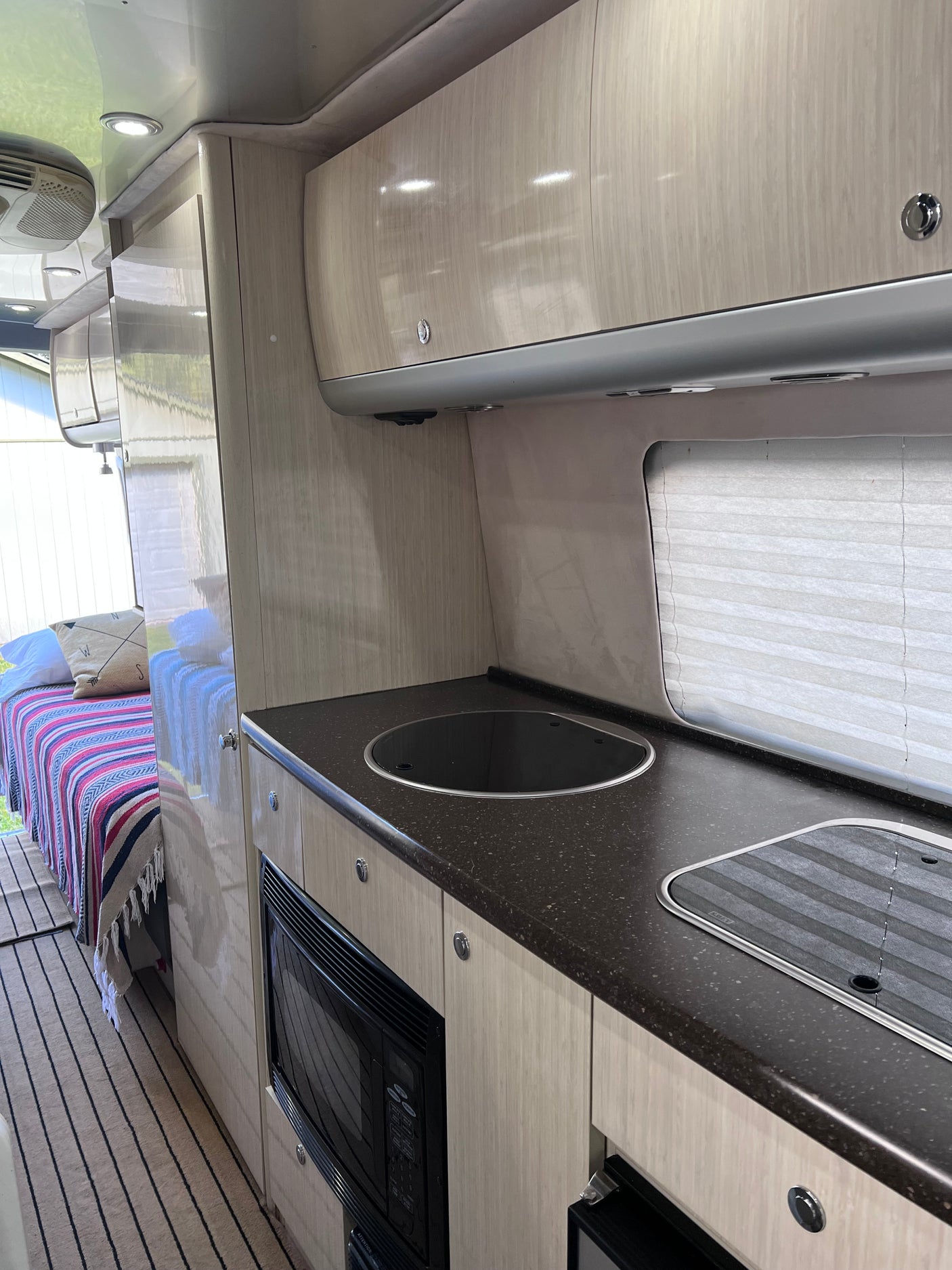 Step inside this RV rental! Modern interior with light wood cabinets, dark countertops, a built-in microwave, and a cozy bed.