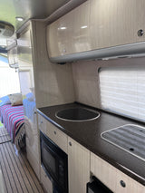 Step inside this RV rental! Modern interior with light wood cabinets, dark countertops, a built-in microwave, and a cozy bed.