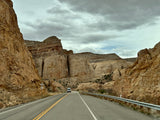 An RV rental van travels a road winding through towering canyon rock formations under a cloudy sky.