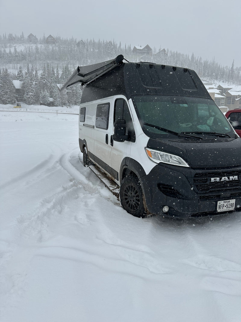 A white Ram ProMaster RV rental in falling snow, parked in a snowy mountain scene with tire tracks leading away. — photo 13