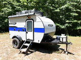 A Sunray Sport compact RV rental, a white and blue camper trailer, parked on dry ground with green trees behind it.