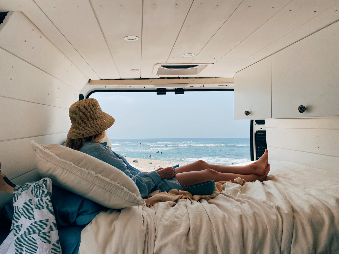 Person in a straw hat relaxes on a bed inside an RV rental, gazing at the ocean and beach scene through the open rear doors.