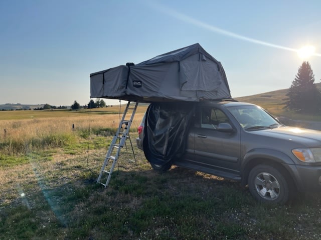  parked on gravel with trees & mountains in background.|This image shows a grey rooftop tent set up on a dark SUV in a sunny — photo 10