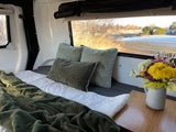 Cozy RV rental interior: a soft bed with green blankets, pillows, and a vibrant flower arrangement by the window overlooking a scenic view.
