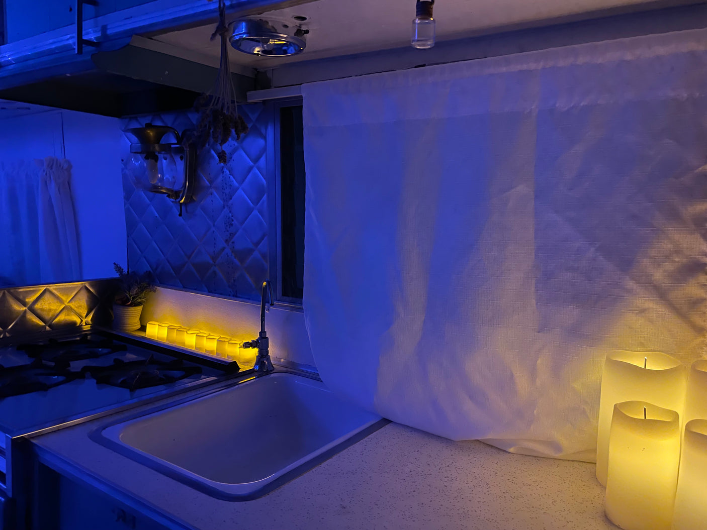 Cozy RV rental kitchen interior at night, with atmospheric blue overhead lighting and warm yellow LED candles illuminating the sink and stove.