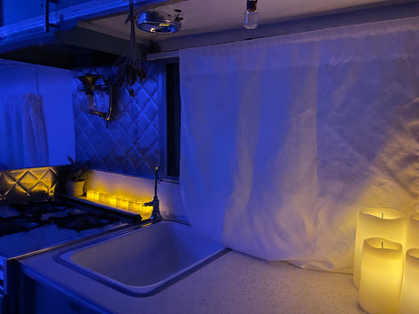 Cozy RV rental kitchen interior at night, with atmospheric blue overhead lighting and warm yellow LED candles illuminating the sink and stove.